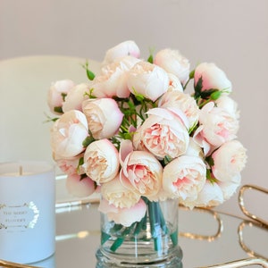 May include: A bouquet of pale pink artificial peonies in a clear glass vase on a gold mirrored tray. The vase has a textured surface. The tray has a gold metal frame.