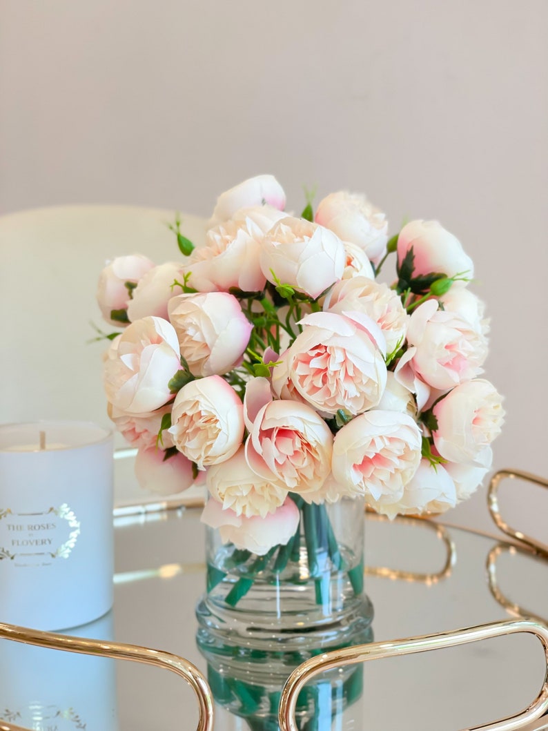May include: A bouquet of pale pink artificial peonies in a clear glass vase on a gold mirrored tray. The vase has a textured surface. The tray has a gold metal frame.