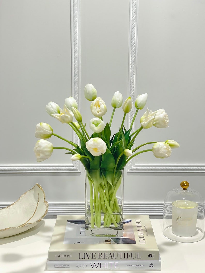 Puede incluir: Un ramo de tulipanes blancos en un jarr&oacute;n de vidrio transparente sobre una mesa con libros. Los libros tienen los t&iacute;tulos "Live Beautiful" y "White".