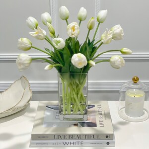 Puede incluir: Un ramo de tulipanes blancos en un jarr&oacute;n de vidrio transparente sobre una mesa blanca con dos libros titulados "Live Beautiful" y "White".