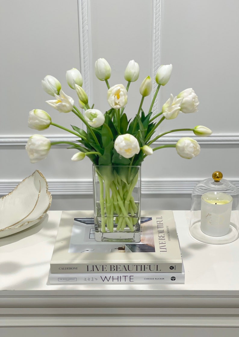 Puede incluir: Un ramo de tulipanes blancos en un jarr&oacute;n de vidrio transparente sobre una mesa blanca con dos libros titulados "Live Beautiful" y "White".