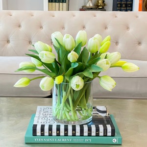 May include: A bouquet of white tulips with green stems in a clear glass vase on a stack of books. The books are titled "Elements of Style", "Elements of Style", and "Yves Saint Laurent The Scandal Collection 1971".