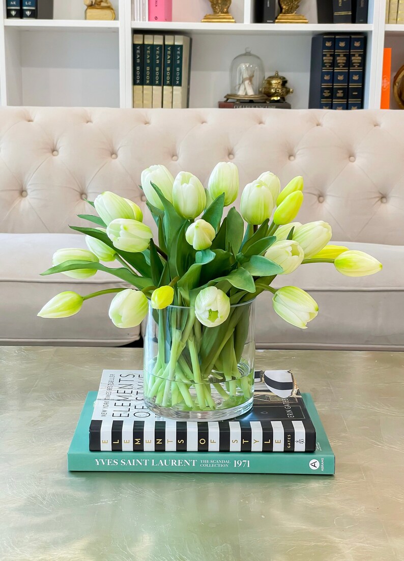 May include: A bouquet of white tulips with green stems in a clear glass vase on a stack of books. The books are titled "Elements of Style", "Elements of Style", and "Yves Saint Laurent The Scandal Collection 1971".