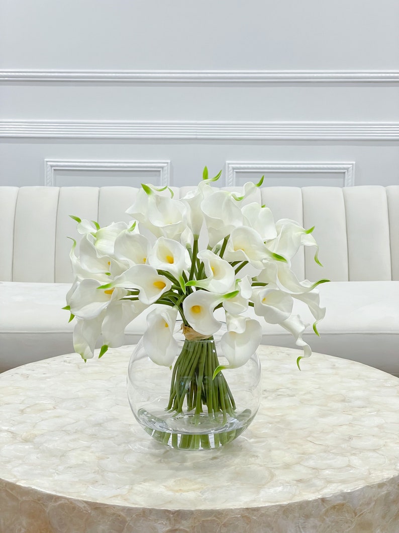 May include: A bouquet of white calla lilies arranged in a clear glass vase on a white table with a white couch in the background.