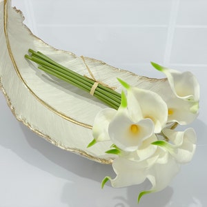 May include: White ceramic feather-shaped tray with a gold rim, holding a bouquet of white calla lilies with green stems.