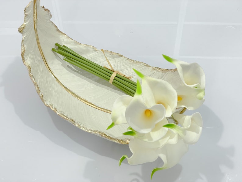 May include: White ceramic feather-shaped tray with a gold rim, holding a bouquet of white calla lilies with green stems.
