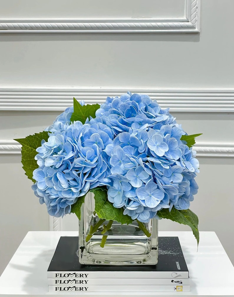 May include: A bouquet of light blue hydrangeas in a clear glass cube vase. The vase is sitting on a stack of magazines with the word "FLOWERY" printed on the covers.