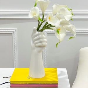 May include: White ceramic vase shaped like a hand holding a bouquet of white calla lilies. The vase is sitting on a stack of books with yellow, pink, and brown covers.