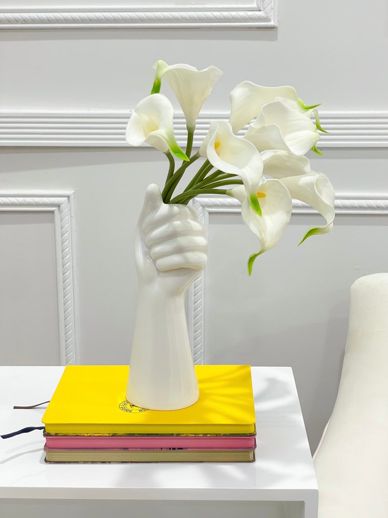May include: White ceramic vase shaped like a hand holding a bouquet of white calla lilies. The vase is sitting on a stack of books with yellow, pink, and brown covers.