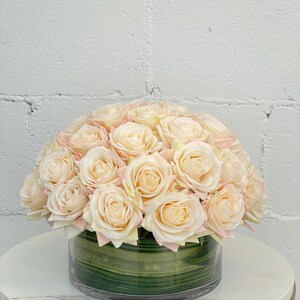 May include: A bouquet of cream-colored artificial roses arranged in a clear glass vase with a green striped liner. The roses are arranged in a dome shape.