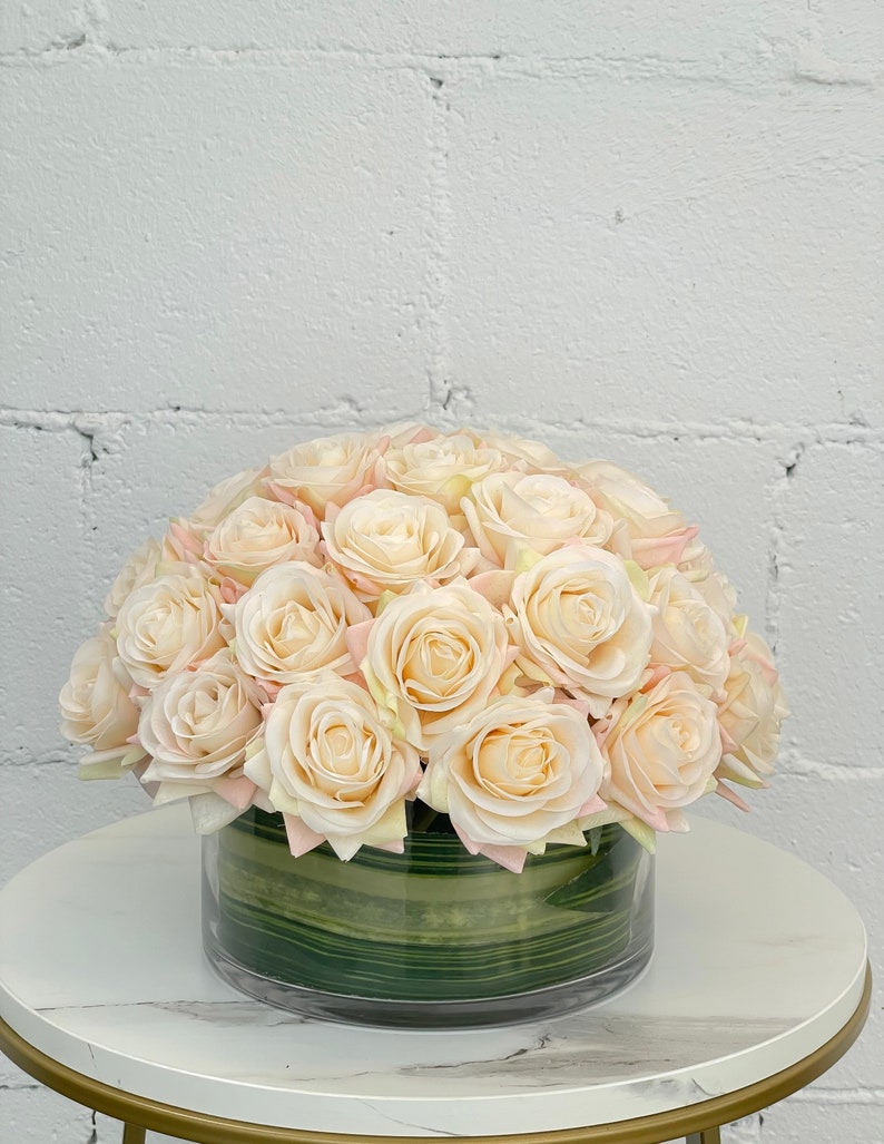 May include: A bouquet of cream-colored artificial roses arranged in a clear glass vase with a green striped liner. The roses are arranged in a dome shape.
