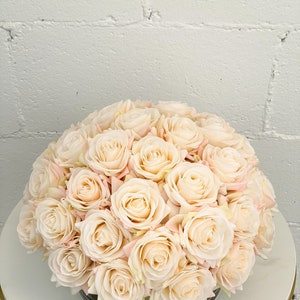 May include: A bouquet of cream-colored artificial roses arranged in a clear glass vase. The roses are clustered together in a circular shape, creating a full and lush appearance.