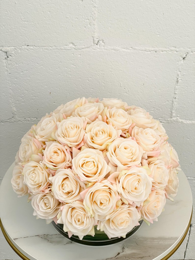 May include: A bouquet of cream-colored artificial roses arranged in a clear glass vase. The roses are clustered together in a circular shape, creating a full and lush appearance.