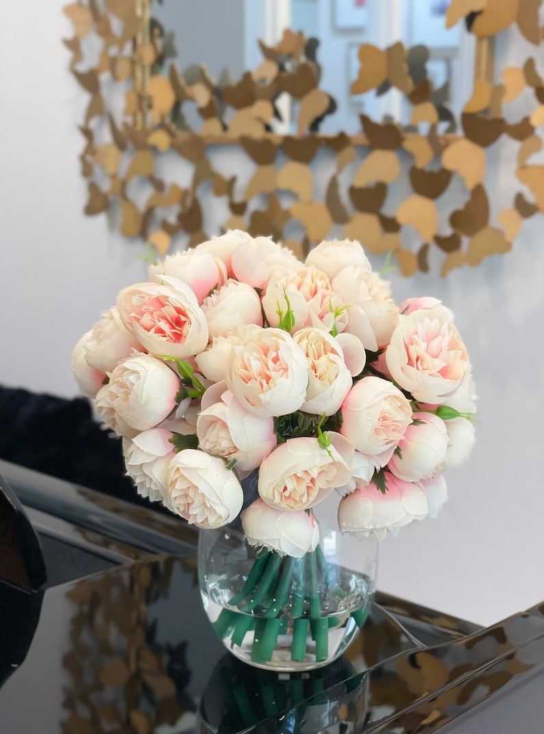 May include: A bouquet of pale pink peonies arranged in a clear glass vase. The vase is sitting on a black surface.