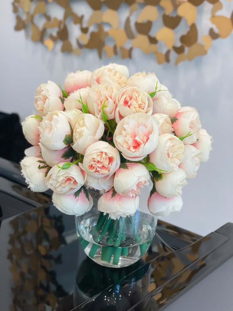 May include: A bouquet of pale pink peonies arranged in a clear glass vase. The vase is sitting on a black surface.