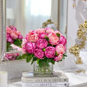 K&ouml;nnte beinhalten: Ein Strau&szlig; rosa Pfingstrosen in einer klaren Glasvase auf einer wei&szlig;en Oberfl&auml;che mit zwei B&uuml;chern mit den Titeln "Live Beautiful" und "White".