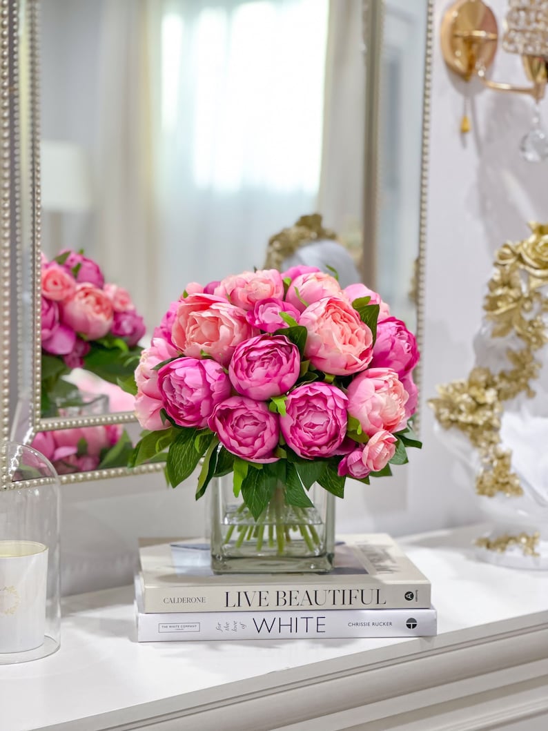 K&ouml;nnte beinhalten: Ein Strau&szlig; rosa Pfingstrosen in einer klaren Glasvase auf einer wei&szlig;en Oberfl&auml;che mit zwei B&uuml;chern mit den Titeln "Live Beautiful" und "White".