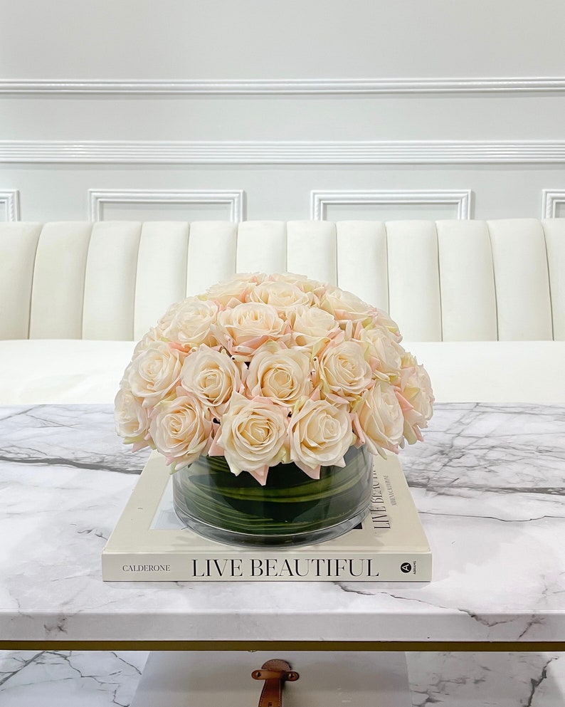 May include: A bouquet of blush pink roses in a clear glass vase with green leaves. The vase is sitting on a white marble coffee table with a book titled "Live Beautiful" by Caldeone.