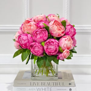 K&ouml;nnte beinhalten: Ein Strau&szlig; rosa Pfingstrosen in einer durchsichtigen Glasvase, die auf zwei B&uuml;chern steht. Das obere Buch tr&auml;gt den Titel "Live Beautiful White" von Chrissie Rucker.