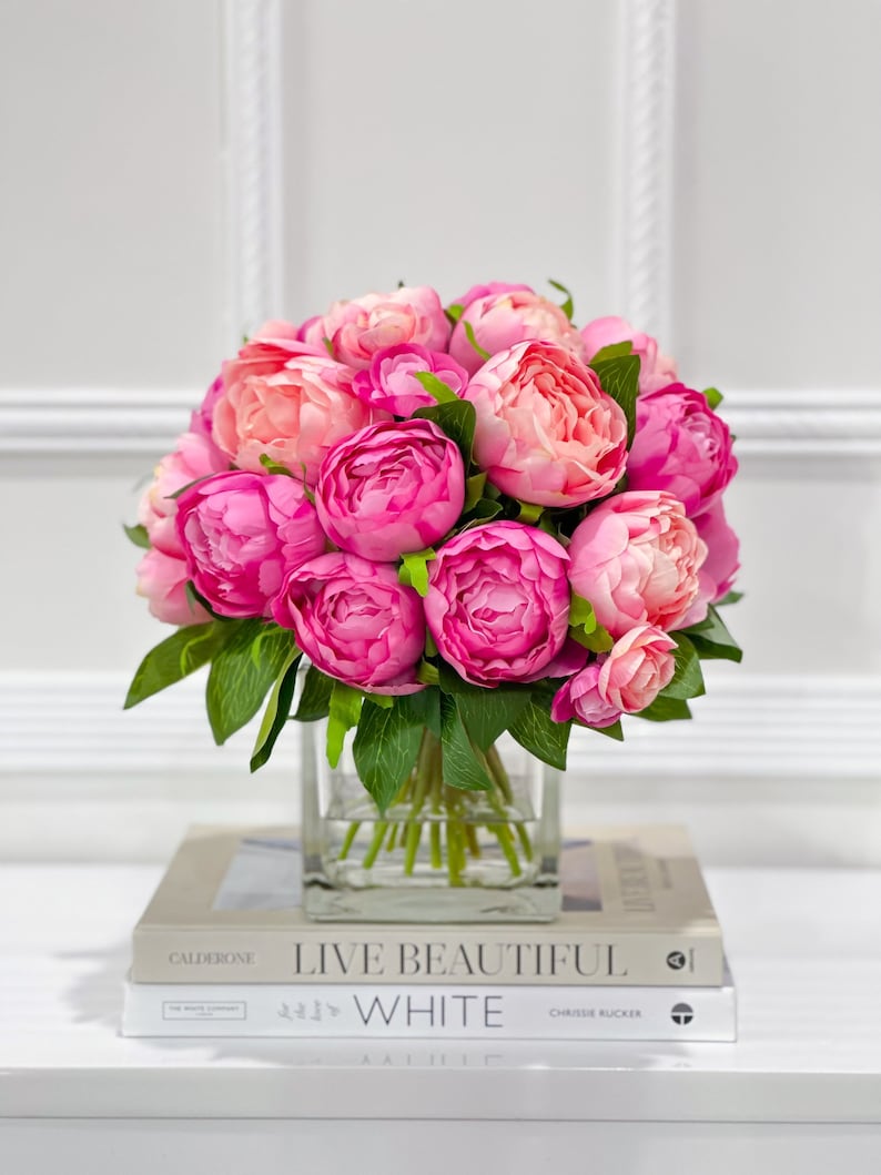 K&ouml;nnte beinhalten: Ein Strau&szlig; rosa Pfingstrosen in einer durchsichtigen Glasvase, die auf zwei B&uuml;chern steht. Das obere Buch tr&auml;gt den Titel "Live Beautiful White" von Chrissie Rucker.