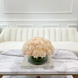 May include: A white sofa with a glass vase filled with a bouquet of blush pink roses on a marble coffee table. The table has a book on it with the text "Live Beautiful".