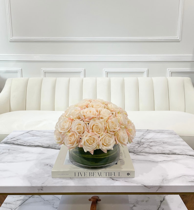 May include: A white sofa with a glass vase filled with a bouquet of blush pink roses on a marble coffee table. The table has a book on it with the text "Live Beautiful".