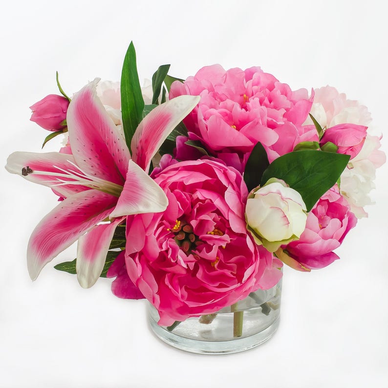 May include: A bouquet of pink peonies and a pink lily in a clear glass vase. The flowers are arranged in a symmetrical pattern and are in full bloom.