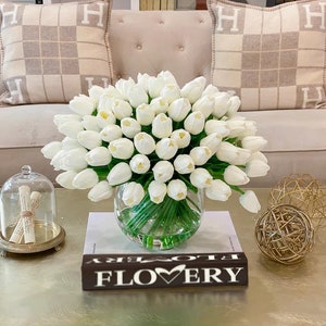 May include: A bouquet of white artificial tulips in a clear glass vase on a coffee table. The vase is sitting on a book with the word "FLOWERY" printed on the cover. There are two gold decorative spheres on the table.