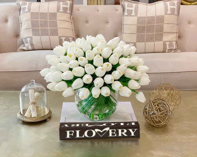 May include: A bouquet of white artificial tulips in a clear glass vase on a coffee table. The vase is sitting on a book with the word "FLOWERY" printed on the cover. There are two gold decorative spheres on the table.