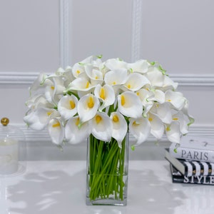 May include: A display of white calla lilies with yellow centres and green stems in a clear, rectangular glass vase. The flowers are arranged on a white surface, with a decorative accent and books in the background.
