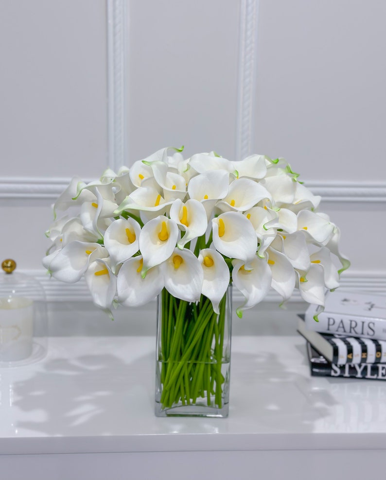 May include: A display of white calla lilies with yellow centres and green stems in a clear, rectangular glass vase. The flowers are arranged on a white surface, with a decorative accent and books in the background.