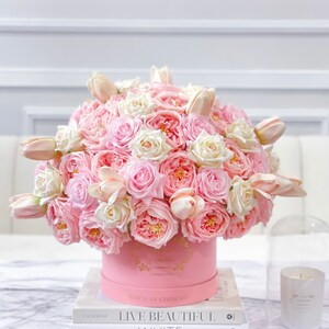 Puede incluir: Un arreglo floral rosa y blanco en una caja de sombrero rosa. La caja tiene letras doradas que dicen "The Roses by Hello". El arreglo est&aacute; sobre una mesa con un libro titulado "Live Beautiful, White" de Chrissie Rucker.