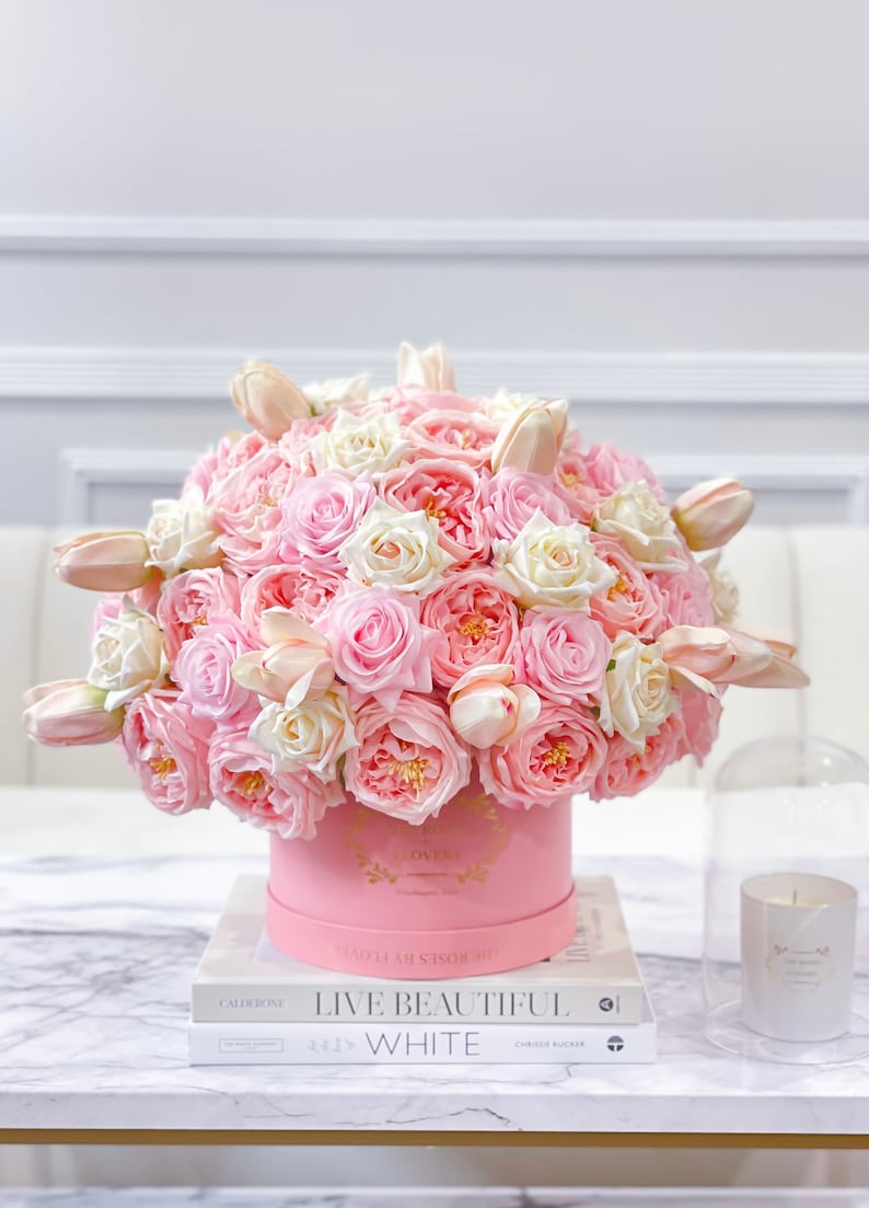 Puede incluir: Un arreglo floral rosa y blanco en una caja de sombrero rosa. La caja tiene letras doradas que dicen "The Roses by Hello". El arreglo est&aacute; sobre una mesa con un libro titulado "Live Beautiful, White" de Chrissie Rucker.