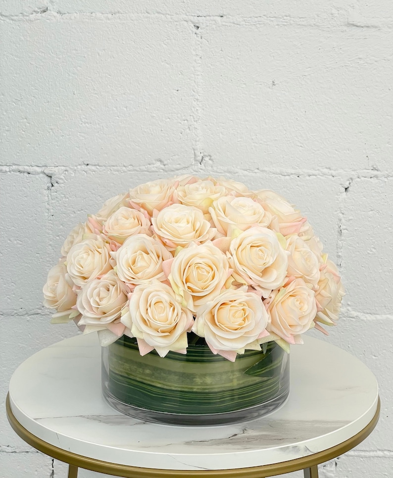 May include: A bouquet of blush pink artificial roses arranged in a clear glass vase with green leaves. The vase is sitting on a white marble table with a gold base.