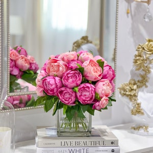 K&ouml;nnte beinhalten: Ein Strau&szlig; rosa Pfingstrosen in einer klaren Glasvase auf einem Stapel B&uuml;cher. Die B&uuml;cher sind mit "Live Beautiful" und "White" betitelt.