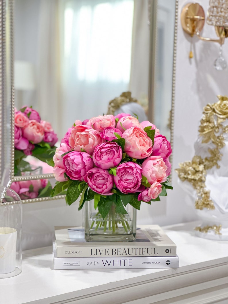 K&ouml;nnte beinhalten: Ein Strau&szlig; rosa Pfingstrosen in einer klaren Glasvase auf einem Stapel B&uuml;cher. Die B&uuml;cher sind mit "Live Beautiful" und "White" betitelt.