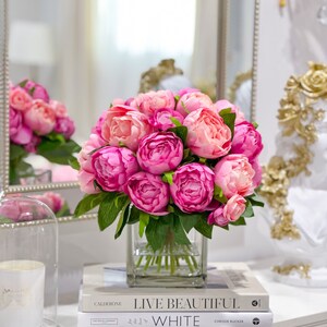 K&ouml;nnte beinhalten: Ein Strau&szlig; rosa Pfingstrosen in einer klaren Glasvase, die auf zwei B&uuml;chern steht. Die B&uuml;cher sind mit "Live Beautiful" und "The White Company" betitelt.