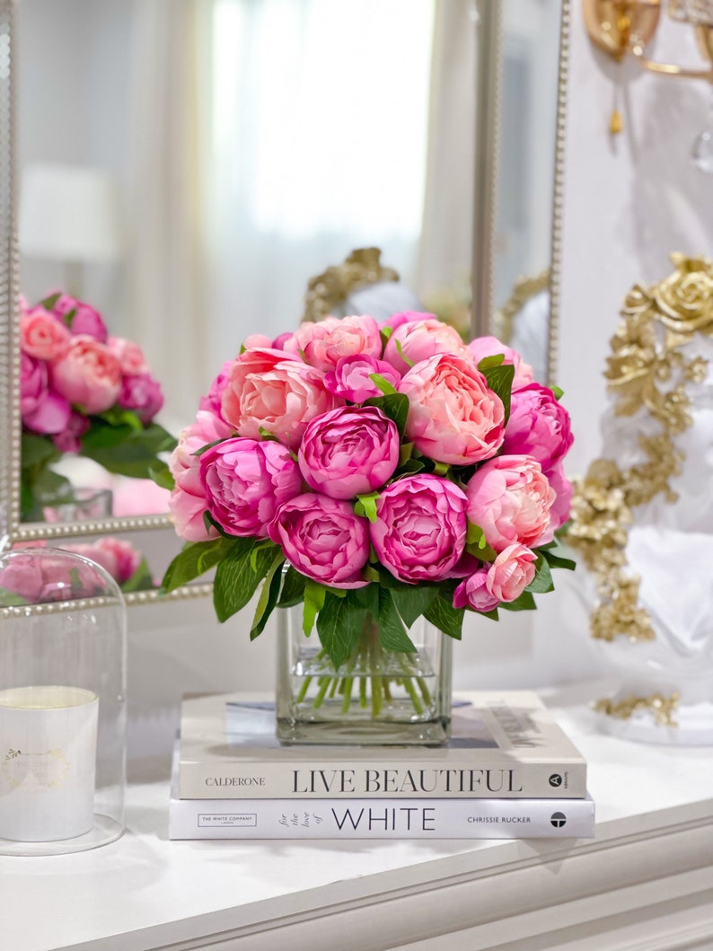 K&ouml;nnte beinhalten: Ein Strau&szlig; rosa Pfingstrosen in einer klaren Glasvase, die auf zwei B&uuml;chern steht. Die B&uuml;cher sind mit "Live Beautiful" und "The White Company" betitelt.