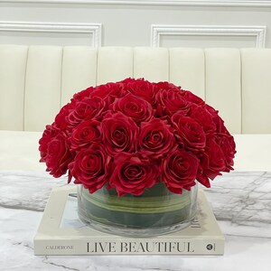May include: A bouquet of red silk roses arranged in a clear glass vase with green accents. The vase is sitting on a white book with the text "LIVE BEAUTIFUL" on the cover.