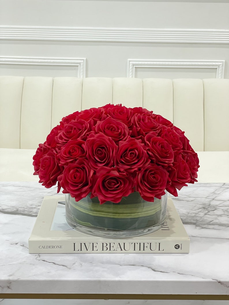 May include: A bouquet of red silk roses arranged in a clear glass vase with green accents. The vase is sitting on a white book with the text "LIVE BEAUTIFUL" on the cover.