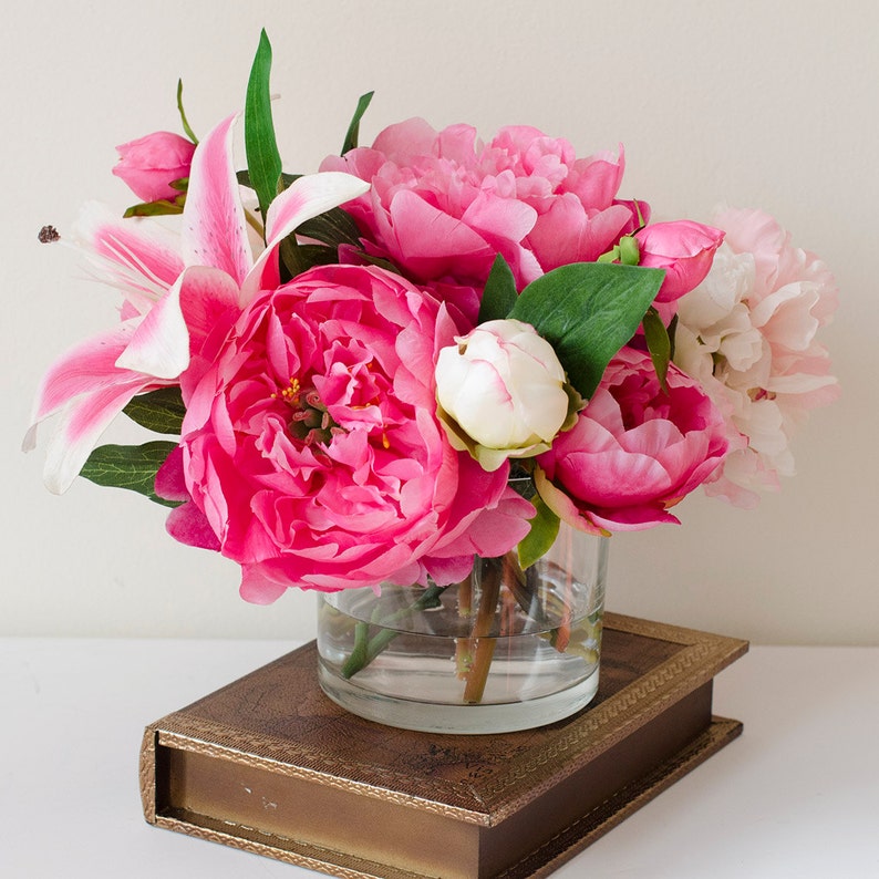 May include: A bouquet of pink peonies and white lilies arranged in a clear glass vase. The vase is sitting on a brown book with gold accents.