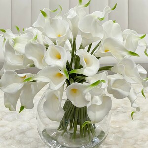 May include: A bouquet of white calla lilies arranged in a clear glass vase. The vase is sitting on a white marble surface.