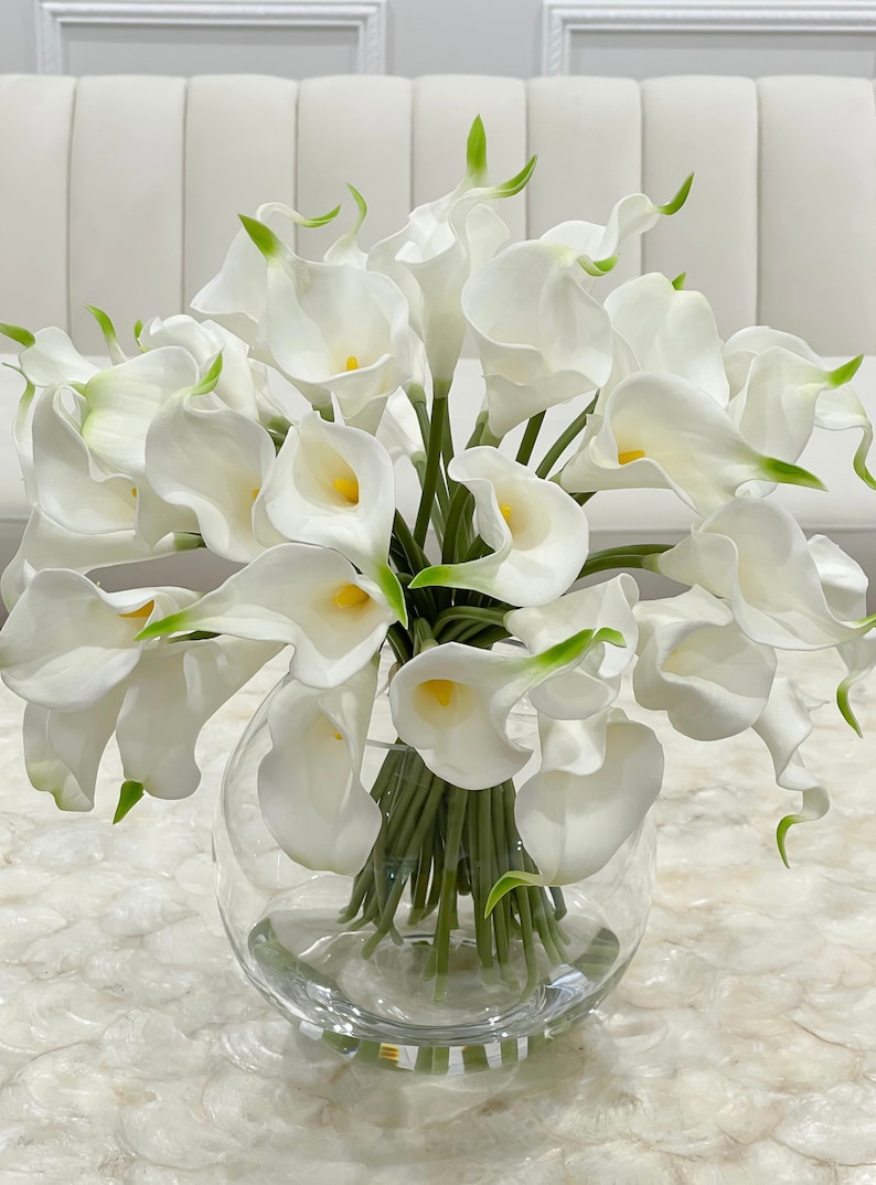 May include: A bouquet of white calla lilies arranged in a clear glass vase. The vase is sitting on a white marble surface.