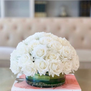 May include: A white rose bouquet in a clear glass vase with a green base. The vase is sitting on a book with the title "VALENTINO THEMES AND VARIATIONS" on the cover.