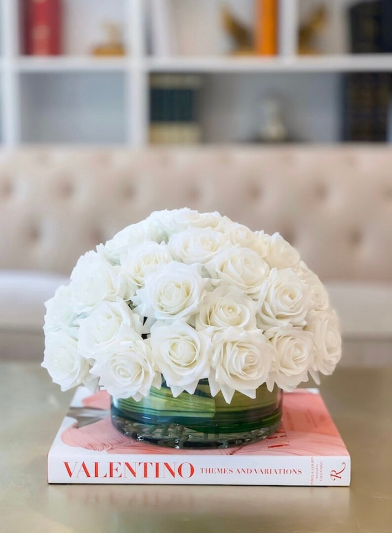 May include: A white rose bouquet in a clear glass vase with a green base. The vase is sitting on a book with the title "VALENTINO THEMES AND VARIATIONS" on the cover.