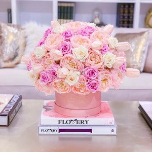 Puede incluir: Una caja de sombrero rosa llena de un ramo de rosas y tulipanes rosas, blancos y melocot&oacute;n. La caja est&aacute; encima de dos libros con la palabra "FLOWERY" impresa en la portada.