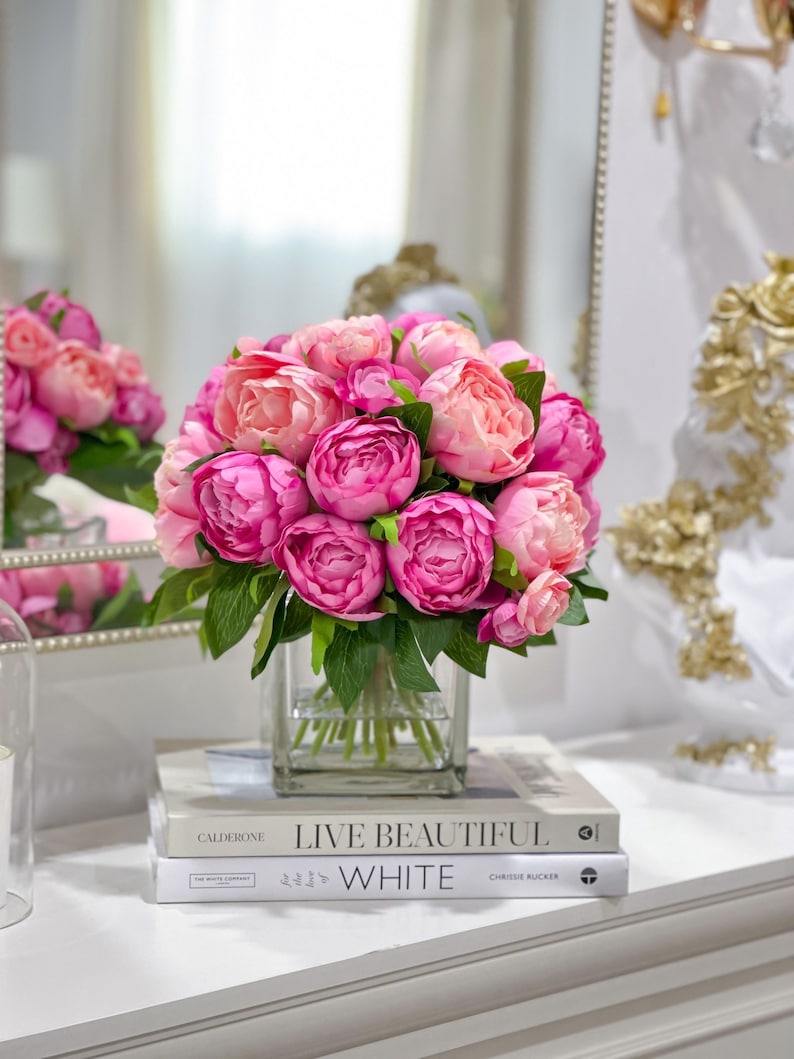 K&ouml;nnte beinhalten: Ein Strau&szlig; rosa Pfingstrosen in einer klaren Glasvase auf einer wei&szlig;en Oberfl&auml;che mit zwei B&uuml;chern, "Live Beautiful" und "The Love of White" von Chrissie Rucker.