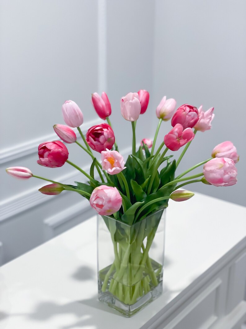 Puede incluir: Un ramo de tulipanes rosas y blancos arreglados en un jarr&oacute;n de vidrio transparente. El jarr&oacute;n est&aacute; sobre una mesa blanca.