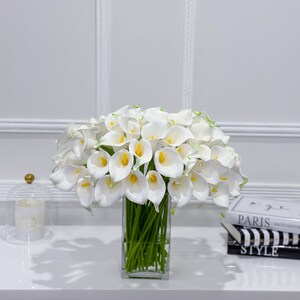 May include: A display of white calla lilies with yellow centres and green stems in a clear, rectangular glass vase. The flowers are densely arranged, creating a full, elegant display. The vase sits on a white surface.