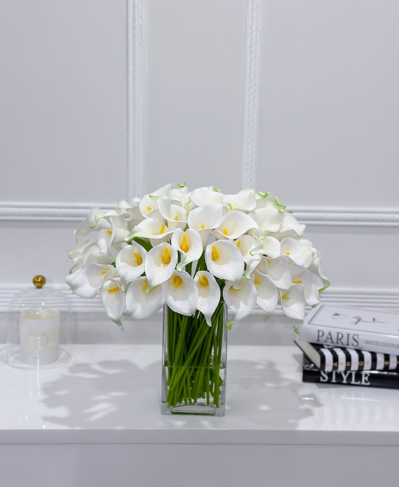 May include: A display of white calla lilies with yellow centres and green stems in a clear, rectangular glass vase. The flowers are densely arranged, creating a full, elegant display. The vase sits on a white surface.